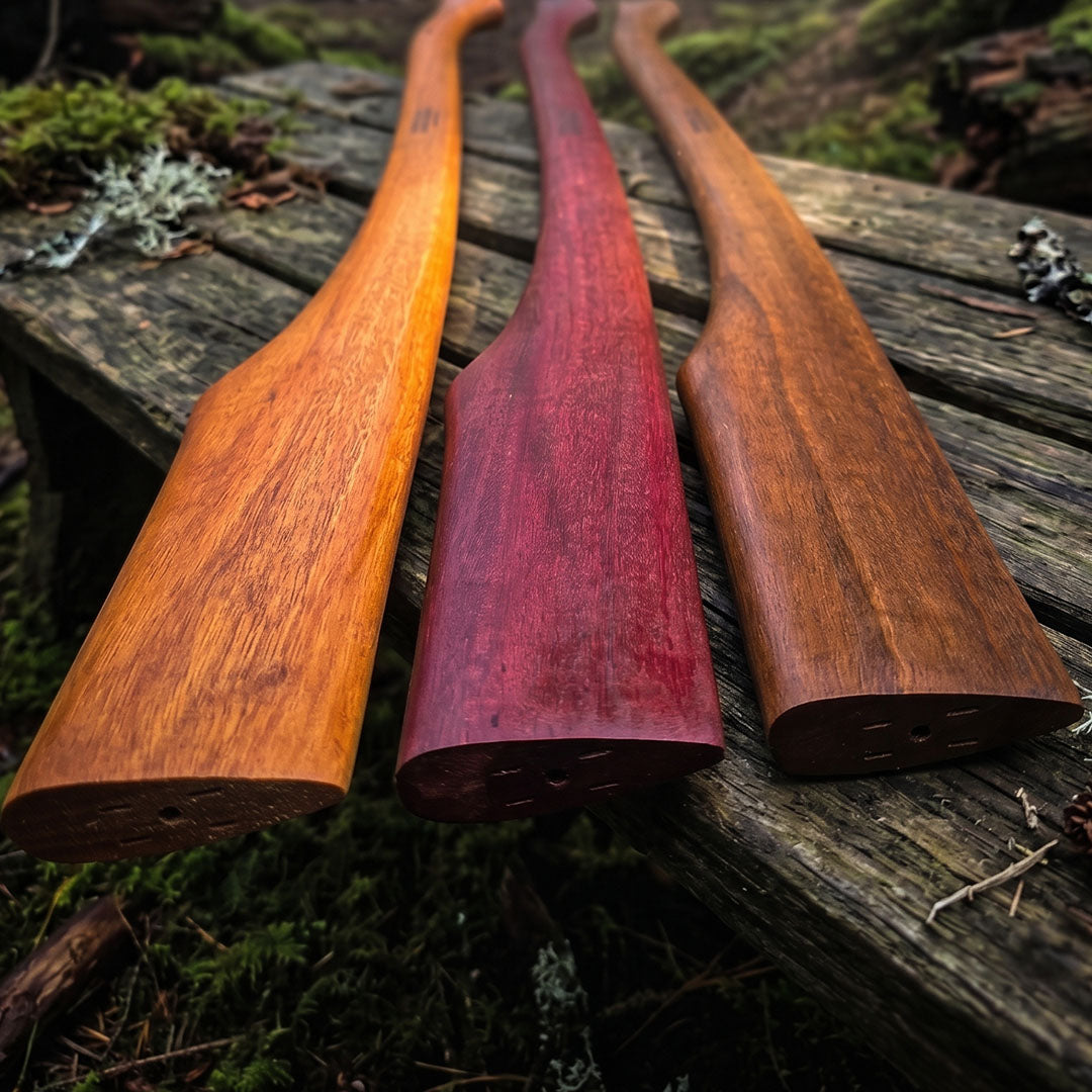Three spotted gum Outbaxe axe handles of different colors on a wooden surface with a natural background.